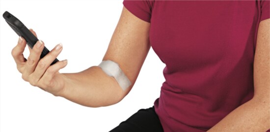 A woman texting on a phone with a piece of gauze on her forearm secured with Nexcare™ Tape.
