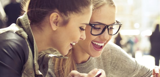 Two smiling, young women looking at a screen with one wearing an unnoticeable clear acne patch.
