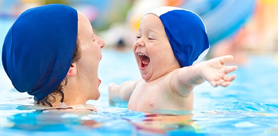 Une femme tient son bébé dans une piscine. Ils portent tous deux des bonnets de bain et se font face.
