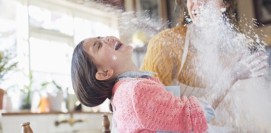 A young girl and her mom laughing, with a cloud of flour in the kitchen.
