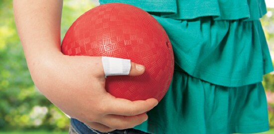 A young girl's hand grasping a dodgeball, with a Nexcare™ Bandage on her thumb.
