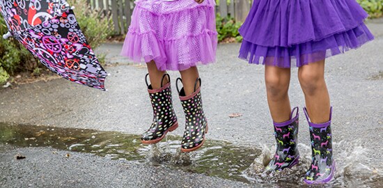 Two girls in rain boots who are jumping in a puddle.
