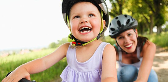 Une jeune fille porte un casque et fait du vélo avec sa mère qui est derrière elle et qui porte aussi un casque.
