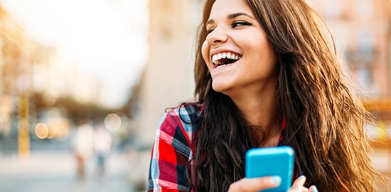 Une jeune femme est penchée sur son téléphone cellulaire dans une rue en pleine ville et rit.
