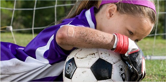 A young girl who is laying down in front of a soccer net, grasping a soccer ball. She is dirty from diving for the ball, and has a Nexcare™ Brand bandage on her elbow.
